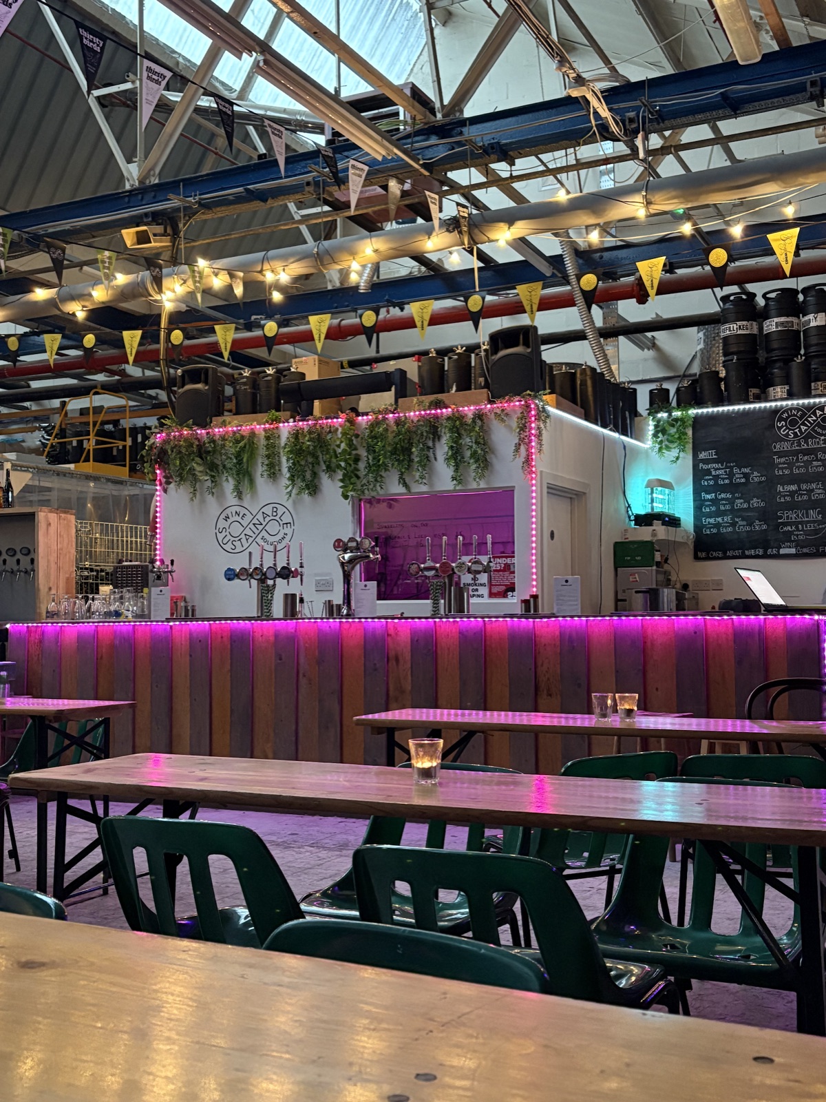 Inside the Borough Wines E17 Taproom, a warehouse space with long communal tables and green chairs under string lights, and the wine-on-tap bar beneath a chalkboard list.